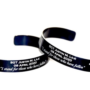 May include: Two black metal bracelets with engraved text and an American flag design. The text reads "SGT JUSTIN M. LILE 09 APRIL 2020 "I stand for those who have fallen"." One bracelet also has a cross engraved on it.