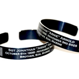May include: Two black metal cuff bracelets with white engraved text. One bracelet features an American flag and the other a Marine Corps emblem. The text includes names, dates, and the word "MARINE".