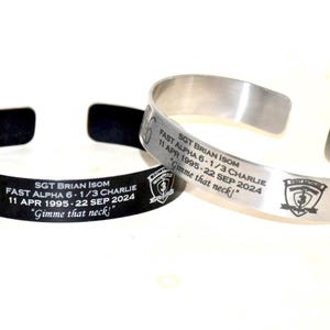 May include: Two stainless steel bracelets with engraved text. The bracelet on the left is black and the bracelet on the right is silver. Both bracelets have the same text: "SGT BRIAN ISOM FAST ALPHA 6-1/3 CHARLIE 11 APR 1995-22 SEP 2024 'Gimme that neck!'".
