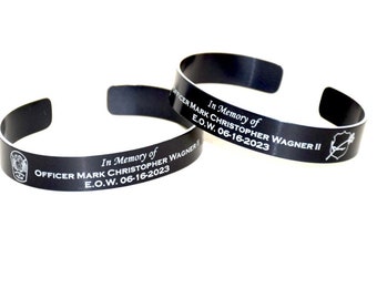 Op de afbeelding: Twee zwarte metalen armbanden met witte gegraveerde tekst. De tekst luidt "In Memoriam van Officer Mark Christopher Wagner II E.O.W. 06-16-2023" en "In Memoriam van Officer Mark Christopher Wagner E.O.W. 06-16-2023".