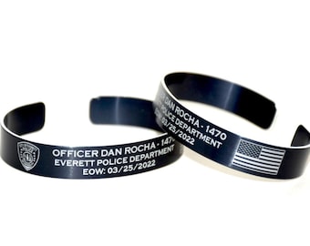 Op de afbeelding: Twee zwarte metalen manchetarmbanden. Eén armband toont de tekst "OFFICER DAN ROCHA - 1470 EVERETT POLICE DEPARTMENT EOW: 03/25/2022" en een politiebadge. De andere armband heeft de tekst en een Amerikaanse vlag.
