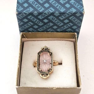 May include: A gold-tone ring featuring an elongated octagonal setting with a pale pink, faceted glass stone and a small clear center accent. The ring is presented in a square box with a blue lid printed with the word AVON repeatedly.