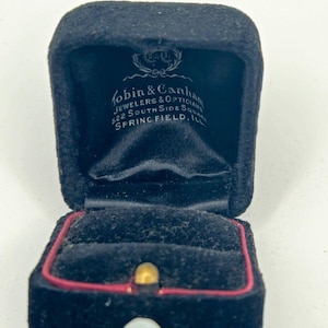 May include: A black velvet ring box with a hinged lid. The interior lid features white printed text reading Tobin & Canham Jewelers & Opticians 522 South Side Square Springfield, Ill. The base has a black velvet insert with a red trim and a white button.