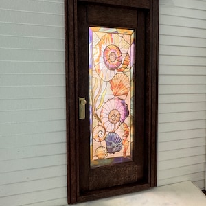 Pode incluir: Uma porta de madeira escura com um painel de vidro emplomado com um desenho de conchas. O vidro é iridescente e mostra tons de roxo, rosa, laranja e amarelo.