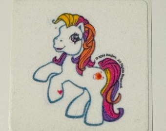 May include: A square sticker featuring a cartoon pony with a white body, blue outlines, and colorful hair. The pony has orange, yellow, pink, and purple hair. A heart is on the pony's flank. The sticker has the text "© 2004 Hasbro, All Rights Reserved."