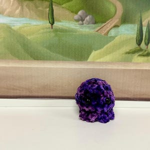 May include: A small, purple and blue plush octopus with black eyes. The octopus is sitting on a white surface.