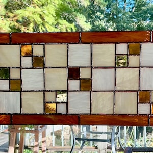 May include: A rectangular stained glass panel featuring a grid of white and cream-colored square tiles interspersed with smaller amber and brown glass accents, all framed by a border of deep orange-brown glass rectangles and small white corners.