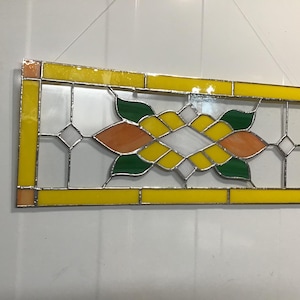May include: Rectangular stained glass panel with a yellow border. The central design features orange, green, white, and yellow floral shapes and diamond accents. The panel is designed to hang and is made with leaded glass.
