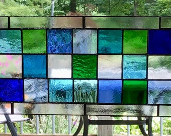 May include: A rectangular stained glass window with a black metal frame. The window features a grid of squares in shades of green, blue, and clear glass. The glass has varying textures and patterns, creating a dynamic visual effect. The window is displayed outdoors.