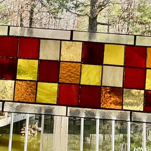 May include: Rectangular stained glass panel with a grid pattern of squares in shades of red, yellow, and amber. The glass has a textured appearance and is framed with a silver-colored metal. The panel is displayed outdoors, with a natural background.