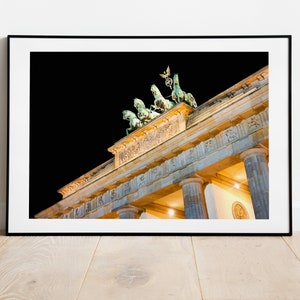 Puede incluir: Una vista nocturna de la Puerta de Brandeburgo en Berl&iacute;n, Alemania. La puerta est&aacute; iluminada con luces amarillas c&aacute;lidas, y los cuatro caballos de bronce y la estatua de la cuadriga en la parte superior son visibles contra el cielo oscuro.