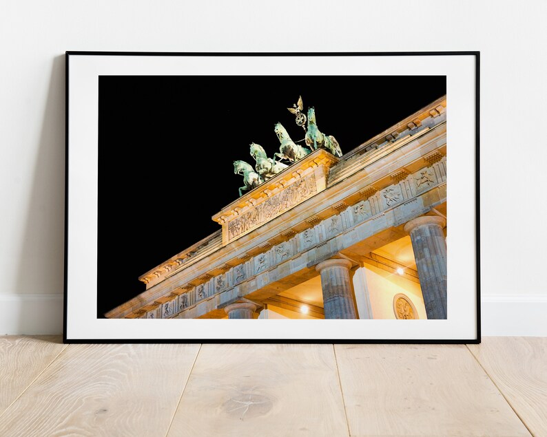 Puede incluir: Una vista nocturna de la Puerta de Brandeburgo en Berl&iacute;n, Alemania. La puerta est&aacute; iluminada con luces amarillas c&aacute;lidas, y los cuatro caballos de bronce y la estatua de la cuadriga en la parte superior son visibles contra el cielo oscuro.
