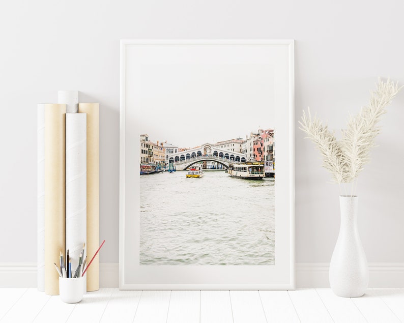 Puede incluir: Una fotograf&iacute;a enmarcada del Puente de Rialto en Venecia, Italia. El puente es blanco con arcos y est&aacute; sobre un canal con barcos. El agua es azul claro y el cielo es gris claro.