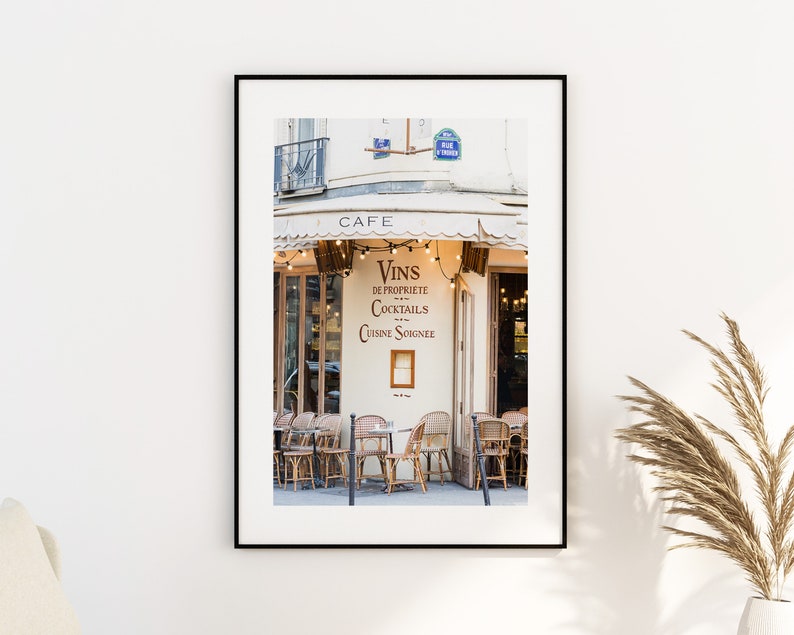 May include: A Parisian cafe with a white awning and a sign that reads "Vins de Proprete Cocktails Cuisine Soignee". The cafe has a few tables and chairs set up outside, and there are string lights hanging above the entrance.