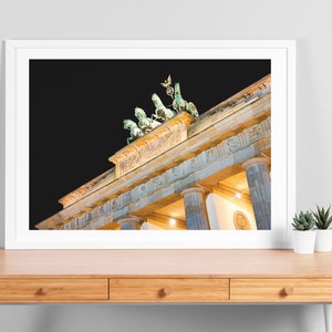Puede incluir: Una vista nocturna de la Puerta de Brandeburgo en Berl&iacute;n, Alemania. La puerta est&aacute; iluminada con luces amarillas c&aacute;lidas, y la estatua de la Cuadriga en la parte superior es visible contra el cielo oscuro.