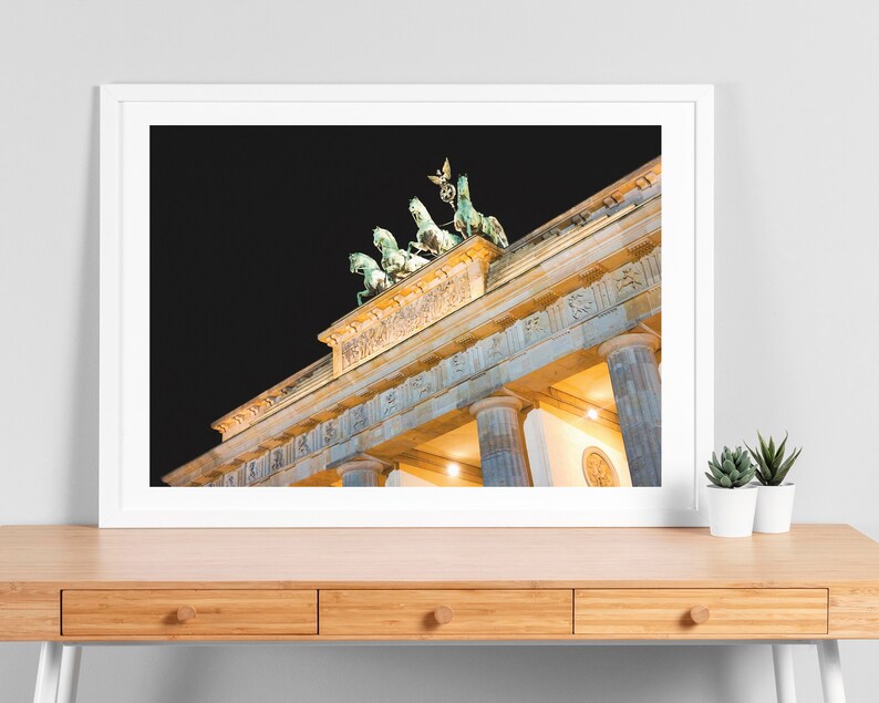 Puede incluir: Una vista nocturna de la Puerta de Brandeburgo en Berl&iacute;n, Alemania. La puerta est&aacute; iluminada con luces amarillas c&aacute;lidas, y la estatua de la Cuadriga en la parte superior es visible contra el cielo oscuro.