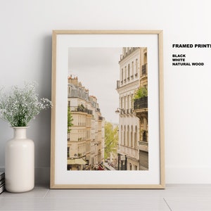 May include: A framed print of a Parisian street scene with buildings in a warm beige color. The buildings have balconies and windows. The print is framed in a natural wood frame.