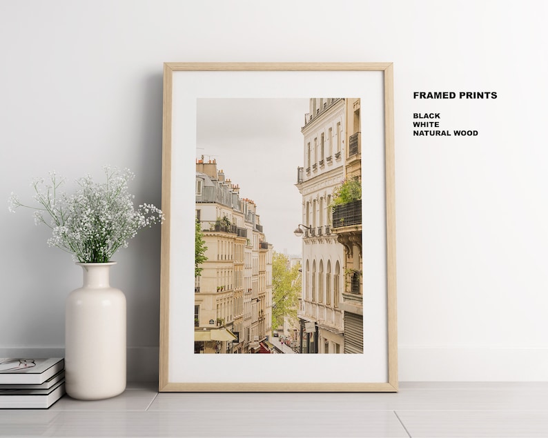May include: A framed print of a Parisian street scene with buildings in a warm beige color. The buildings have balconies and windows. The print is framed in a natural wood frame.