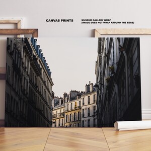 May include: A black and white photograph of a narrow street in Paris, France. The street is lined with tall buildings with many windows. The buildings are old and have a worn look. The street is empty and the sky is overcast.