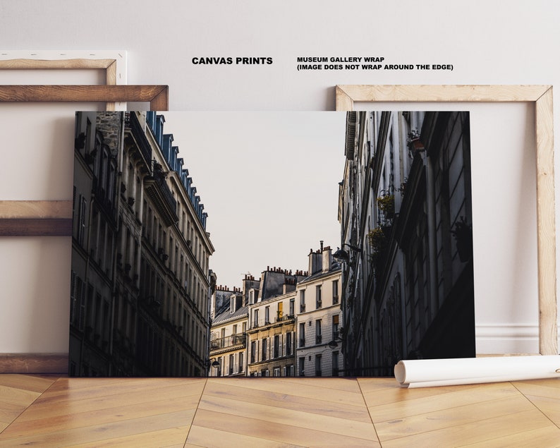 May include: A black and white photograph of a narrow street in Paris, France. The street is lined with tall buildings with many windows. The buildings are old and have a worn look. The street is empty and the sky is overcast.