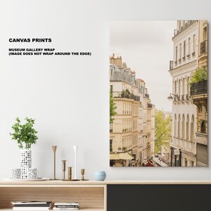 May include: A canvas print of a narrow street in Paris, France. The buildings are white and have balconies. The street is lined with trees and there are people walking on the sidewalk.