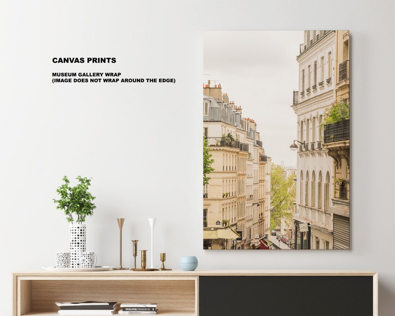 May include: A canvas print of a narrow street in Paris, France. The buildings are white and have balconies. The street is lined with trees and there are people walking on the sidewalk.