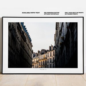 May include: A black and white photograph of a narrow street in Paris, France. The street is lined with tall buildings with windows and balconies. The buildings are in shadow, but the street is lit by the sun. The text "PARIS" is printed at the bottom of the image.