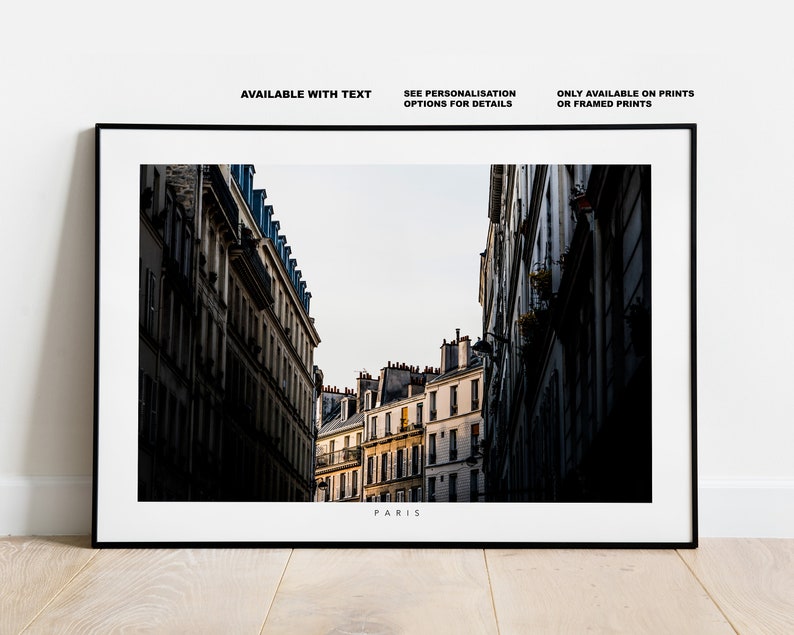 May include: A black and white photograph of a narrow street in Paris, France. The street is lined with tall buildings with windows and balconies. The buildings are in shadow, but the street is lit by the sun. The text "PARIS" is printed at the bottom of the image.