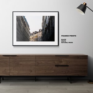 May include: A black framed print of a city street with tall buildings. The print is hanging on a white wall above a wooden cabinet. A black floor lamp with a black shade is on the right side of the image. The text "FRAMED PRINTS" is written in black on the white wall, with the words "BLACK", "WHITE", and "NATURAL WOOD" listed below.