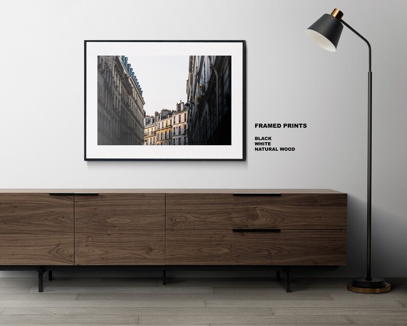 May include: A black framed print of a city street with tall buildings. The print is hanging on a white wall above a wooden cabinet. A black floor lamp with a black shade is on the right side of the image. The text "FRAMED PRINTS" is written in black on the white wall, with the words "BLACK", "WHITE", and "NATURAL WOOD" listed below.
