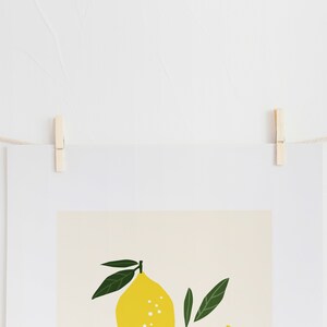 May include: A white framed print with a yellow lemon and green leaves on a beige background. The print is hanging on a white wall with a string and wooden clothespins.