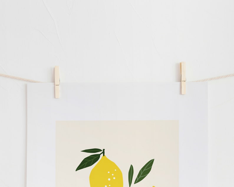 May include: A white framed print with a yellow lemon and green leaves on a beige background. The print is hanging on a white wall with a string and wooden clothespins.
