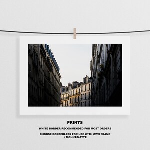 May include: A black and white photograph of a narrow street in Paris, France. The street is lined with tall buildings with windows and balconies. The buildings are in shadow, but the sky is bright and sunny.