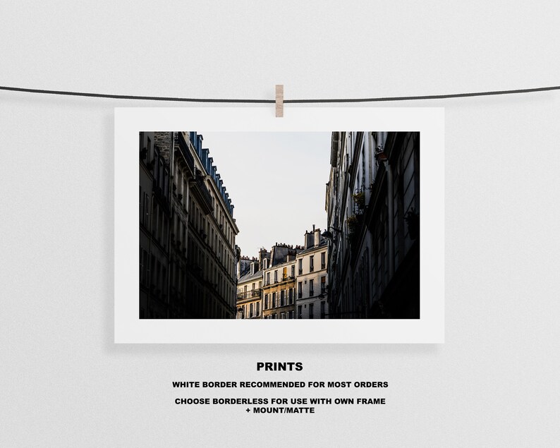 May include: A black and white photograph of a narrow street in Paris, France. The street is lined with tall buildings with windows and balconies. The buildings are in shadow, but the sky is bright and sunny.