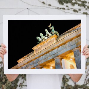Puede incluir: Una fotograf&iacute;a enmarcada de la Puerta de Brandeburgo en Berl&iacute;n, Alemania, de noche. La puerta est&aacute; iluminada con luces amarillas c&aacute;lidas, y el cielo est&aacute; oscuro. La parte superior de la puerta es visible, incluida la escultura de la Quadriga.