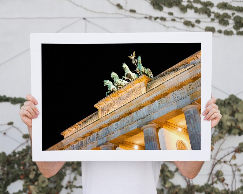 Puede incluir: Una fotograf&iacute;a enmarcada de la Puerta de Brandeburgo en Berl&iacute;n, Alemania, de noche. La puerta est&aacute; iluminada con luces amarillas c&aacute;lidas, y el cielo est&aacute; oscuro. La parte superior de la puerta es visible, incluida la escultura de la Quadriga.