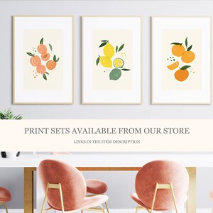 May include: Three framed prints with illustrations of fruit. The first print features peaches, the second print features lemons and limes, and the third print features oranges. The prints are displayed on a white wall above a white table with pink chairs.