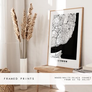 May include: Framed print of a black and white map of Lisbon, Portugal. The map is in a wooden frame with a white mat. The print is on display on a wooden shelf with a vase of dried pampas grass.