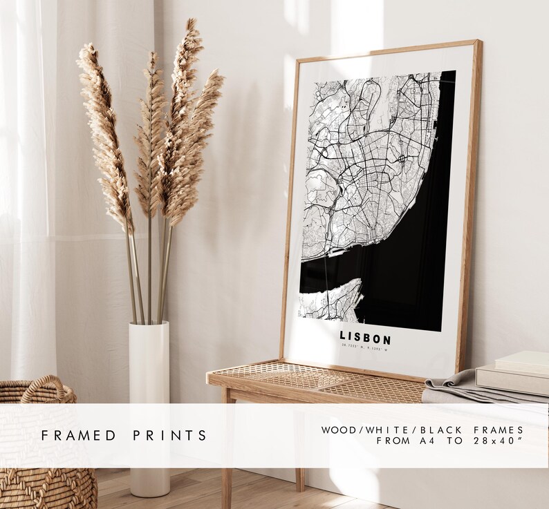 May include: Framed print of a black and white map of Lisbon, Portugal. The map is in a wooden frame with a white mat. The print is on display on a wooden shelf with a vase of dried pampas grass.