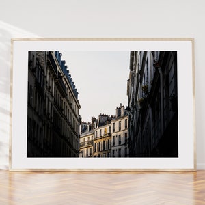 May include: A black and white photograph of a narrow street in Paris, France. The street is lined with tall buildings with many windows. The buildings are in shadow, but the sky is bright.