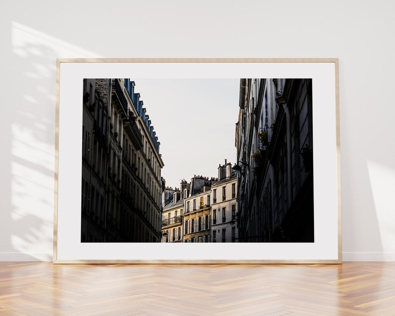 May include: A black and white photograph of a narrow street in Paris, France. The street is lined with tall buildings with many windows. The buildings are in shadow, but the sky is bright.