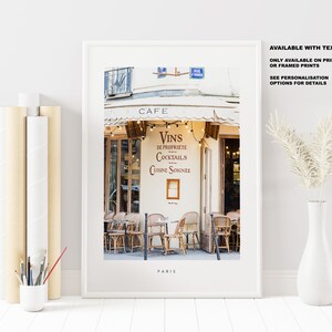 May include: A framed print of a Parisian cafe with a white awning and a sign that reads "Cafe, Vins de Propriete, Cocktails, Cuisine Soignee". The cafe has a few tables and chairs outside, and the street is cobblestone. The print is titled "Paris".