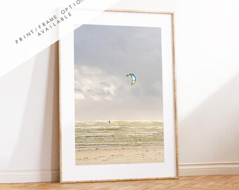 May include: A framed photograph of a kitesurfer on a beach with a blue and white kite in the air. The sky is a light blue with white clouds. The water is a light blue and green.