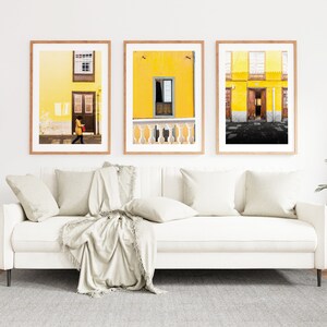 May include: Three framed photographs of yellow buildings with wooden doors and windows. The photos are hanging on a white wall above a white sofa with beige cushions and a throw blanket.