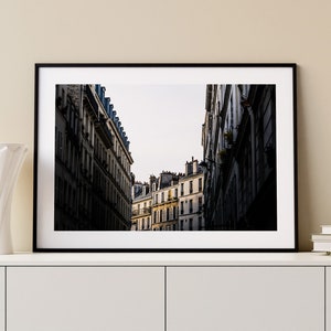 May include: A black and white photograph of a narrow street in Paris, France. The street is lined with tall buildings with many windows. The buildings are in focus, while the street is blurred.