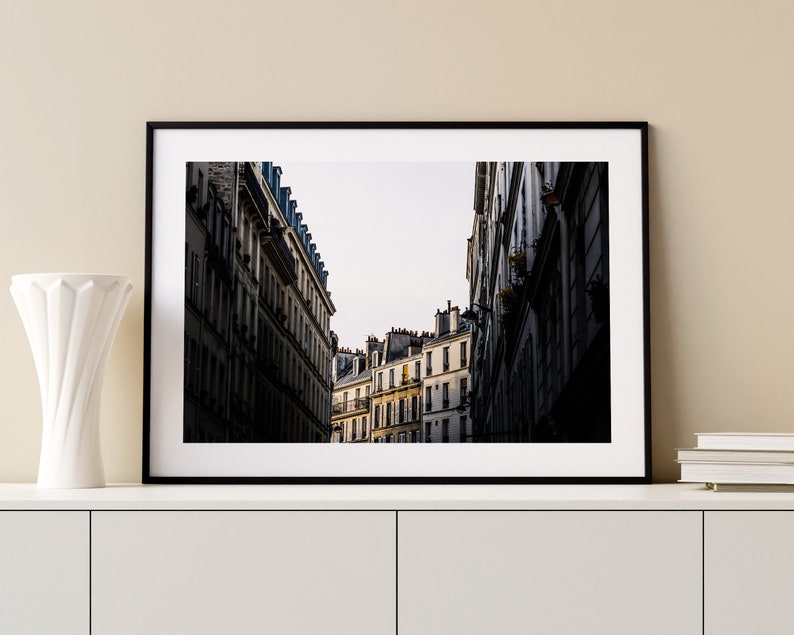 May include: A black and white photograph of a narrow street in Paris, France. The street is lined with tall buildings with many windows. The buildings are in focus, while the street is blurred.