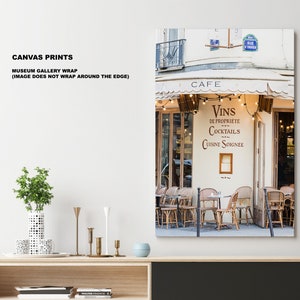 May include: A Parisian cafe with a white awning and a sign that reads "Cafe" and "Vins de Propriete Cocktails Cuisine Soignee". The cafe has a string of lights hanging above the entrance and tables and chairs are set up outside.