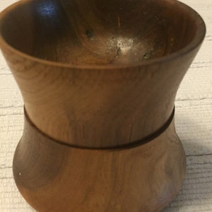 May include: A handcrafted wooden bowl with a unique hourglass shape. The bowl is made of a light brown wood with visible grain patterns. The design features a wide top and base, connected by a narrower midsection. This decorative bowl is suitable for various uses.