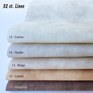 Op de afbeelding: Een stapel van vijf gevouwen stukken 32 ct. linnen stof in diverse neutrale tinten. De kleuren zijn gelabeld als 15 - Creme, 14 - Oyster, 13 - Beige, 12 - Camel en 11 - Hazelnut, wat een verloop toont van licht gebroken wit naar donkerbruin.