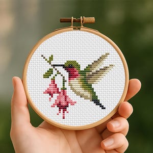 Puede incluir: Un bastidor de bordado circular de madera que contiene un patrón de punto de cruz sobre tela blanca. El diseño presenta un colibrí con plumaje verde y rojo, flotando cerca de un tallo con dos flores fucsia rosas colgantes, en puntos pixelados.